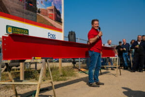 Man in red Vaderstad shirt and blue jeans speaking through microphone in front of red beam and project sign in background.