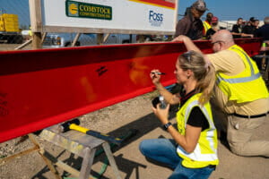 Woman and man in safety yellow vest kneeling down to sign their name on a red construction beam.