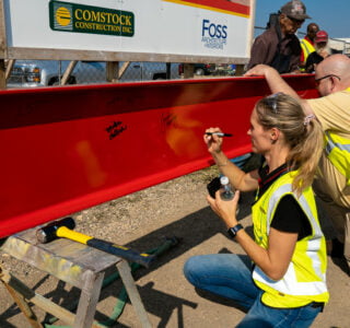 Woman and man in safety yellow vest kneeling down to sign their name on a red construction beam.